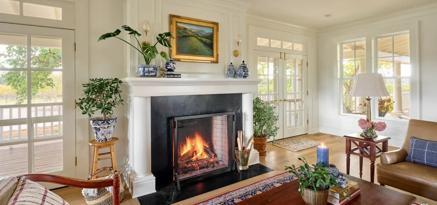 Interior remodel featuring a double-sided fireplace with a traditional mantel, surrounded by historic-style white trim. The space is flanked by two sets of classic French doors that allow natural light to pour in, blending timeless architectural details with modern functionality.