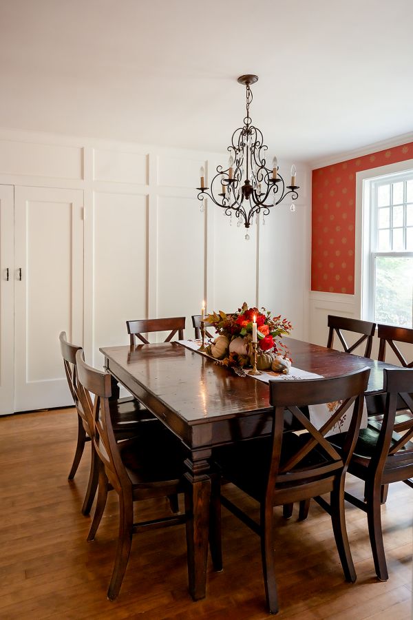 Overall view of dining room with historic wainscoting and wood trim, coral hand-stamped wallpaper, and a large square table featuring a fall centerpiece, under a black and crystal chandelier.