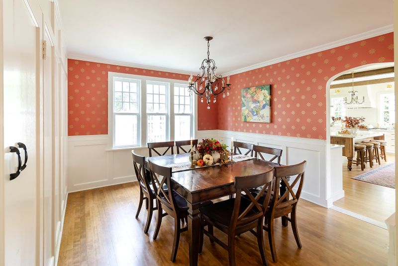 Dining room with historic wainscoting, coral hand-stamped wallpaper, and a large square table with fall centerpiece, with light streaming through three windows.