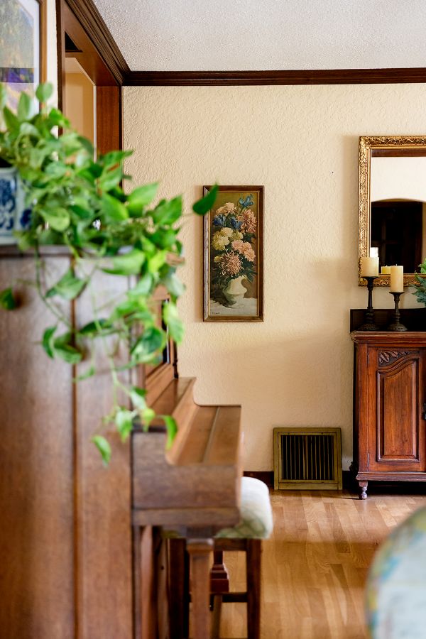 Traditional living room with upright piano and trailing green plant, vintage floral wall art, and wooden cabinet with mirror and candles.