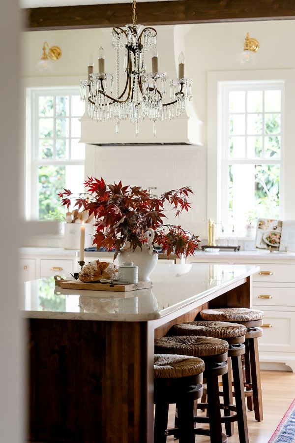 Bright kitchen with a large island, quartz countertop, woven barstools, crystal chandelier, and autumn leaves arranged in a vase, with natural light streaming through tall windows.