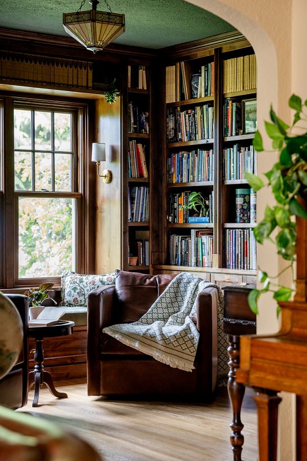 Study with custom built-in bookcase, leather armchair with blanket, large window with garden view, and cozy reading nook.