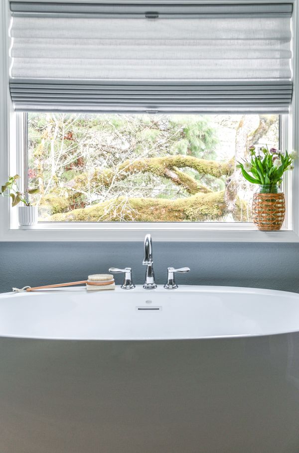 Straight on view of free-standing tub positioned beneath window with honeycomb window blinds.