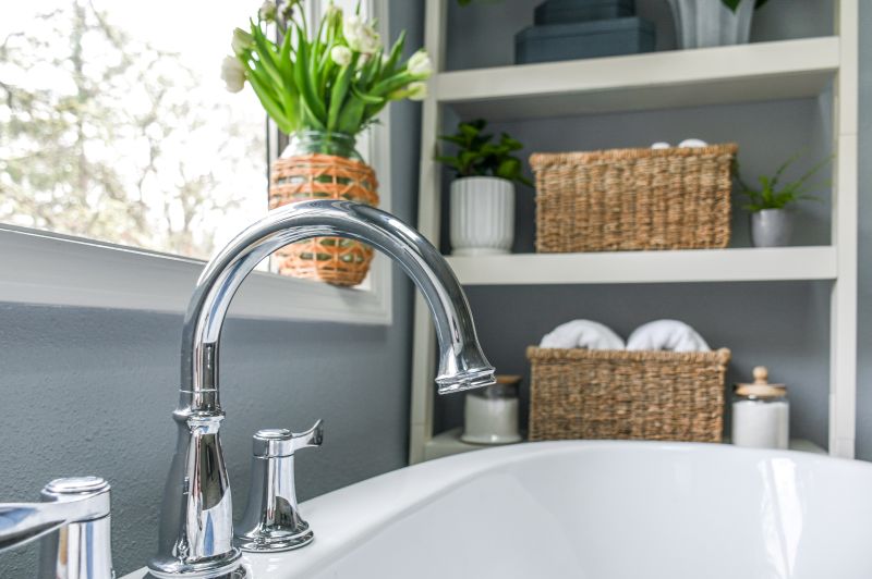 Brilliant polished chrome tub filler over free-standing bathtub with white shelving in background to hold all bathtub essentials.